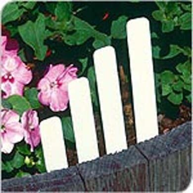 Four white rectangular plant markers of varying heights are stuck in soil near pink flowers and green leaves, bordered by a wooden edge.