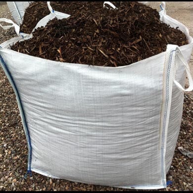 A large white woven bag filled with dark brown mulch sits on a gravel surface. The bag has handles on each corner for lifting and is open at the top, showing the mulch inside.