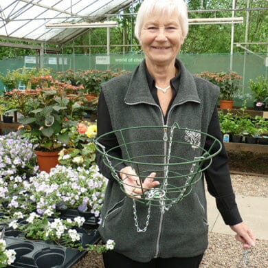 Smiling older woman with short white hair holds an empty wire hanging basket in a greenhouse, surrounded by potted flowering plants and gardening supplies.