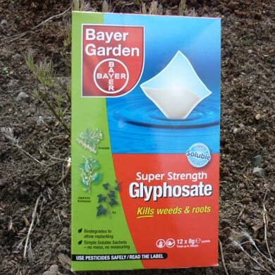 A Bayer Garden box of Super Strength Glyphosate, featuring a white sachet and images of weeds. Text highlights that it kills weeds and roots. The background shows soil and plants.